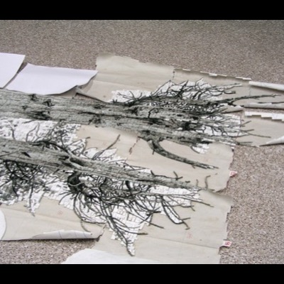 Overhead photograph of a large glass-tile mosaic panel of tree bark and branches laid flat on a studio floor, surrounded by torn paper templates and loose tile scraps.