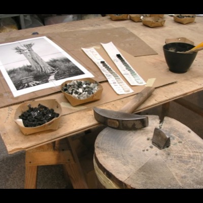 Photograph of a mosaic artist’s worktable: a black-and-white reference photograph of the champion cedar propped up beside strips of graduated grey and white glass tile samples, trays of cut tesserae, and a hammer and hardie for cutting tile.