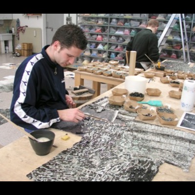 Photograph of two mosaic artists at a long worktable covered in trays of colored glass tesserae, one cutting tile with a hardie and hammer while assembling a grey-toned mosaic panel of the tree.