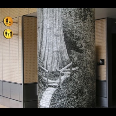 Closer photograph of the same "Champion" column beside the airport restroom entrance, framing the mosaic’s boardwalk and cedar trunk against the perforated wall panel and accessible-restroom icon.