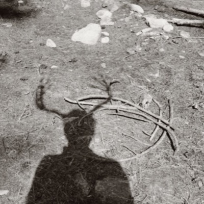 Black-and-white photograph of a photographer’s own shadow with arms raised like branching antlers, cast across a rocky, sunlit slope of grass and stone.