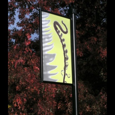 Enamel way-finding sign mounted on a black post beside a residential street, showing a black-and-white fern fiddlehead on yellow with autumn trees and a passing Metro bus behind it.