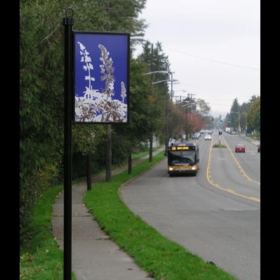 Enamel way-finding sign mounted on a black post along a sidewalk, showing the black-and-white lupine image on blue, with a Metro bus signed "120 White Center" passing on the street behind.