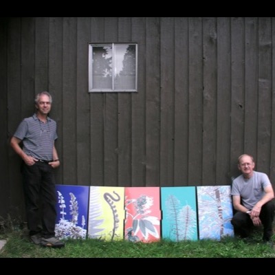 Photograph of two men posing beside all five "Flora" enamel panels leaned against a dark wood-sided building, one standing at left and one crouching at right.