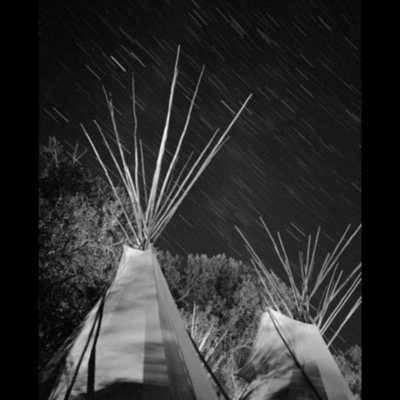 Black-and-white photograph of a man in ceremonial dress with a feather headdress and painted face, sitting cross-legged in front of a tipi holding a beaded pipe bag and feathered items.