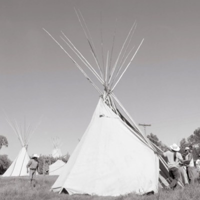 Black-and-white photograph, silhouetted at dusk, of a tipi’s bundled smoke-flap poles crossing in front of a crescent moon.