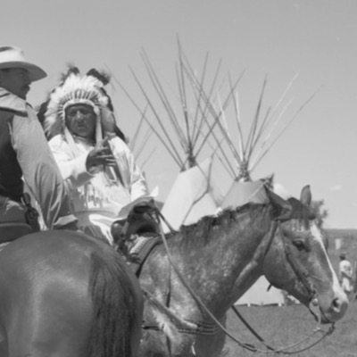 Black-and-white portrait of a man in a full eagle-feather headdress and fringed regalia, seated cross-legged in front of a tipi holding a beaded pipe bag and eagle-wing fan, face marked with paint.