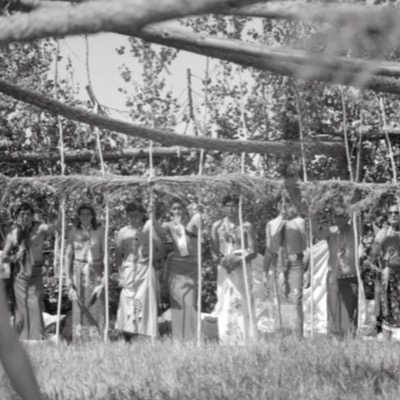 Black-and-white photograph, seen through overhead branches, of eight people standing beneath a brush arbor holding vertical poles, most bare-chested and wrapped in cloth for a ceremony.