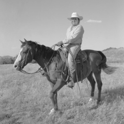 Black-and-white photograph of a man in a light-colored hat and jeans, smiling atop a saddled horse in an open pasture with hills behind.