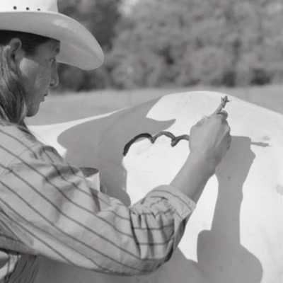 Black-and-white photograph, from behind, of a woman in a straw hat painting a wavy black line onto a large white surface with a stick, a paint can in her other hand.