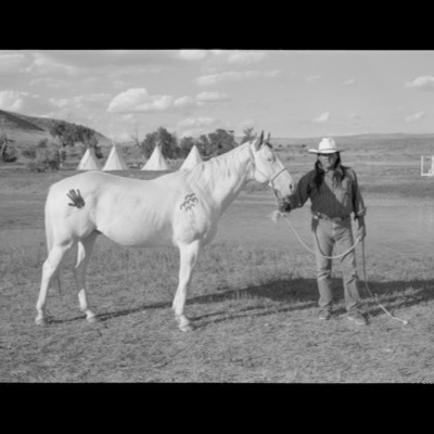 Black-and-white photograph of a man in a cowboy hat standing beside a white horse painted with a black handprint and wavy line on its flank, tipis lining the water’s edge behind them.