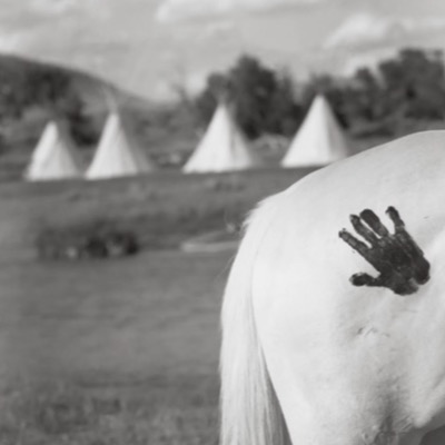 Black-and-white close-up photograph of a black handprint painted on the hindquarters of a white horse, tipis visible blurred in the background.