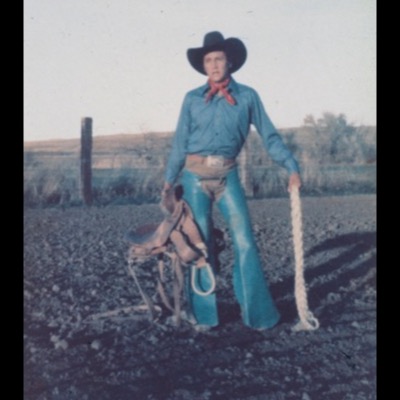 Color archival snapshot (credited "Photo Real Bird Family, 1970’s," not a Peter de Lory photograph) of a young man in a turquoise western outfit and black hat posing with a saddle and coiled rope in a gravel yard.