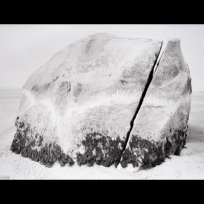 Peter de Lory, "Rock Harbor Rock," Cape Cod, Massachusetts, 1973 — a black-and-white photograph of a large, weathered boulder split by a vertical crack, seaweed clinging to its base on a sandy beach.