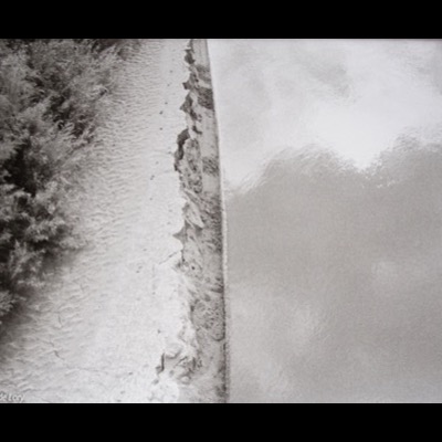 Peter de Lory, "Colorado River Shore," Utah, 1971 — a black-and-white photograph looking down at a riverbank’s edge, sandy shore on the left and rippled water on the right divided by a jagged seam of dark silt.