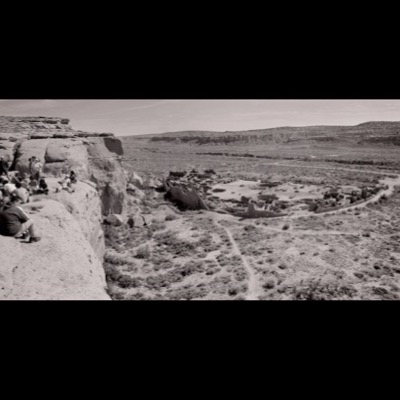 Black-and-white panoramic photograph of a tour group seated and standing on a canyon-rim overlook at Chaco Canyon, the excavated great-house ruins spread across the valley floor below.