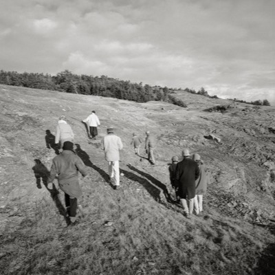Black-and-white photograph of a small group hiking up a windswept, grassy hillside toward a treeline, their long shadows cast across the slope.