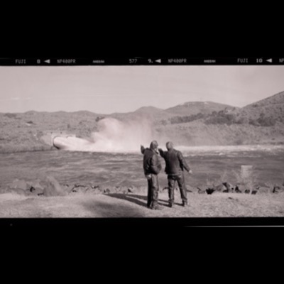 Black-and-white photograph of two men standing at a riverbank pointing toward a dam’s spillway discharging a heavy plume of water and spray into the river below.