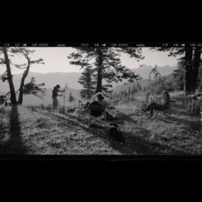 Black-and-white panoramic photograph of a group relaxing at a mountain campsite at dusk, two people seated in camp chairs by a fire while a third tends a sapling, distant peaks visible through the trees.