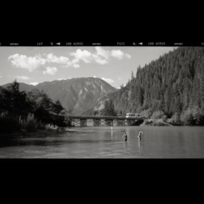 Black-and-white photograph of a person and dog standing at the edge of a bridge over a forested river valley, a motorhome crossing in the background and swimmers wading in the water below.