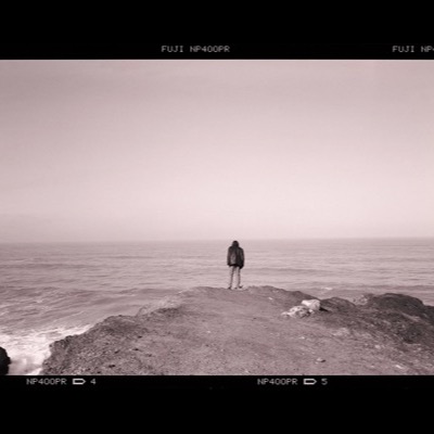 Black-and-white photograph of a lone figure standing at the edge of a foggy sea cliff, back to the camera, looking out over calm water.