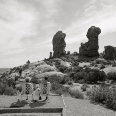 Black-and-white photograph of a figure silhouetted against a rugged, snow-capped mountain range, arms raised, viewed from behind in an open backcountry meadow.