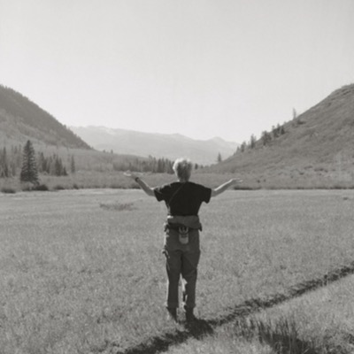 Black-and-white photograph of a person standing at an easel painting a canyon landscape from a snowy overlook, brushes and painting supplies laid out on a nearby table.