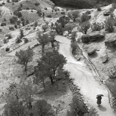 Black-and-white photograph, from inside a natural double-arch formation, of a small child standing far below with arms raised amid scattered boulders.