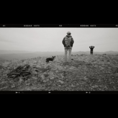 Black-and-white photograph of a lone hiker walking away down a winding dirt road through open, hazy rangeland hills.