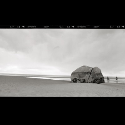 Black-and-white panoramic photograph of a large split boulder resting alone on a wide tidal beach, two figures walking past it under a heavy, overcast sky.