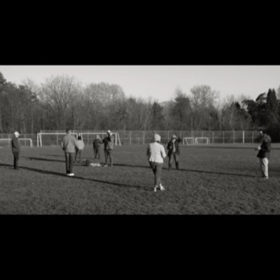 Black-and-white panoramic photograph of a small group standing on a soccer field looking up and photographing the sky, goalposts and a tree line behind them.