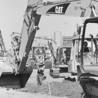 Black-and-white photograph of an elevated guideway under construction beside a rain-slicked highway, blurred cars passing below a crane lifting a girder segment into place.