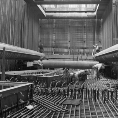 Four black-and-white photographs of an elevated station’s escalators at different construction stages: bare concrete stringers, finished stairs mid-installation, escalators wrapped in plastic, and the completed escalators in use by passengers.