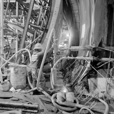 Black-and-white photograph, from below, of a guideway segment being lifted by an overhead gantry crane, two workers in harnesses guiding the concrete piece into position.