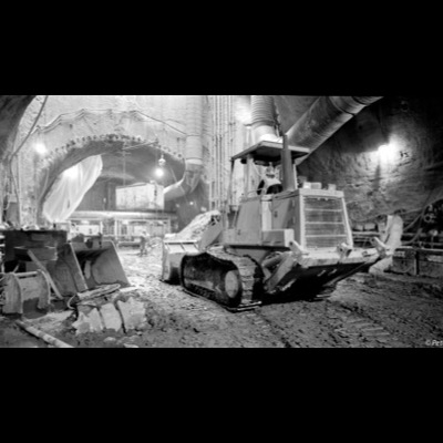 Black-and-white photograph of two ironworkers climbing rebar-caged concrete columns via suspended platforms, densely tied reinforcing steel filling the frame.