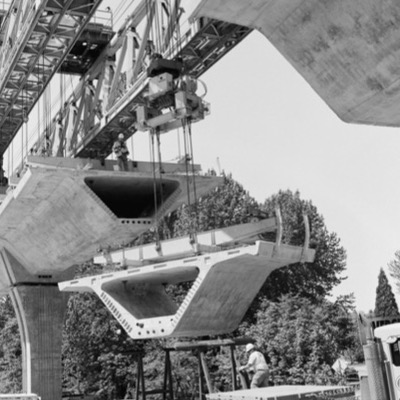 Black-and-white photograph, aerial, of a light rail station and elevated guideway construction site spreading across a wide urban lot, cranes and a high-rise hotel visible in the distance.