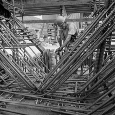 Black-and-white photograph, aerial, of a crane lowering a large object at a light rail construction site, station platforms and columns rising amid excavated ground.