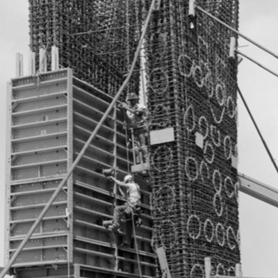 Black-and-white photograph of a large fabric-wrapped and netted object -- likely a rail vehicle component -- suspended from a crane above a wet loading dock, workers in rain gear steadying it below.