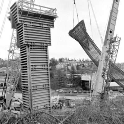 Black-and-white photograph of massive concrete guideway pylons under construction beside a lifted steel-mesh form, cranes and residential hillside visible in the background.