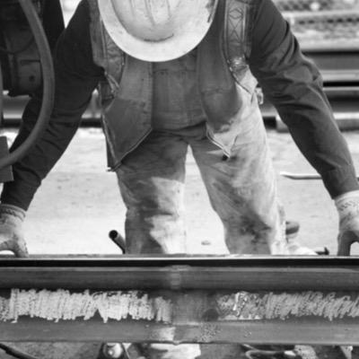 Black-and-white photograph, close up, of a hard-hatted worker in overalls bending to inspect a length of rail resting on the ground.