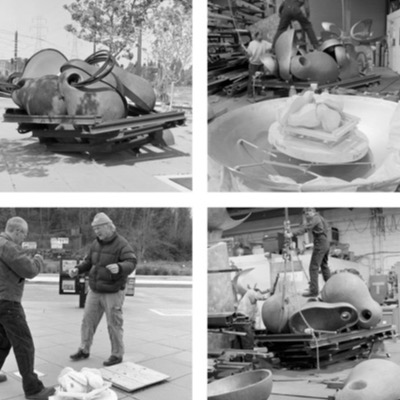 Black-and-white photograph of two men standing over a small model or maquette of the sculpture set on a marked turntable outdoors, gesturing as they discuss its orientation.