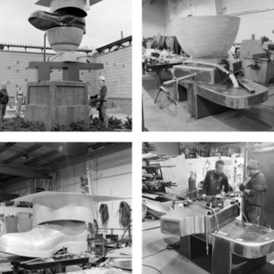 Black-and-white photograph inside the fabrication shop of workers using a crane to lift and position large curved bronze sculpture components onto a rail-mounted base.