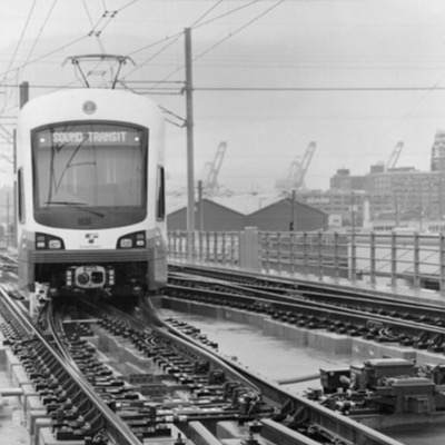 Black-and-white photograph, aerial, of a light rail construction corridor showing excavation, elevated structure, and a distant high-rise hotel across a wide urban site.