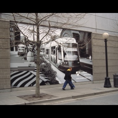 Color photograph of black-and-white construction photographs printed on banners and hung along a chain-link fence beside a downtown street, showing rail-laying work and a repair crew.