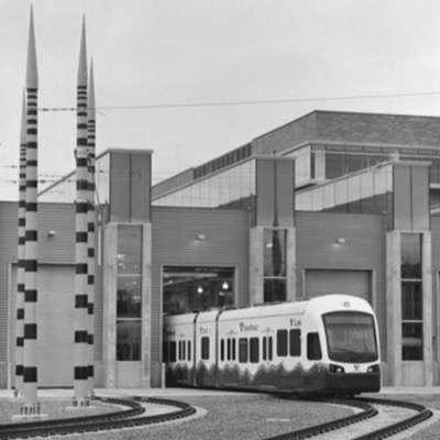 Black-and-white photograph of a light rail train emerging from a striped-pillar maintenance facility, curved track and building visible in the yard.