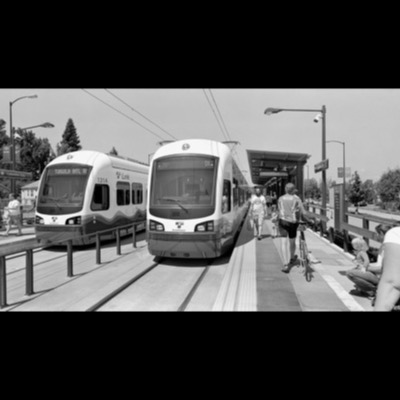 Black-and-white photograph of two Sound Transit light rail trains stopped at the Columbia City station platform, passengers walking past including a cyclist with his bike.