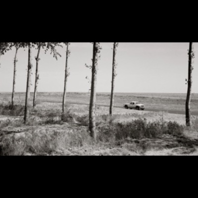 Black-and-white photograph taken through a row of roadside trees of a white pickup truck driving along a straight rural highway across flat, open farmland.