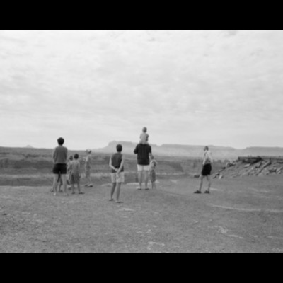 Black-and-white photograph, from behind, of a family group standing at a desert overlook watching the distant mesas and canyon of a high desert landscape.