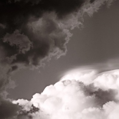 Black-and-white photograph of a dark storm cloud mass split by a brilliant sunlit cumulus tower pushing up through the gap.