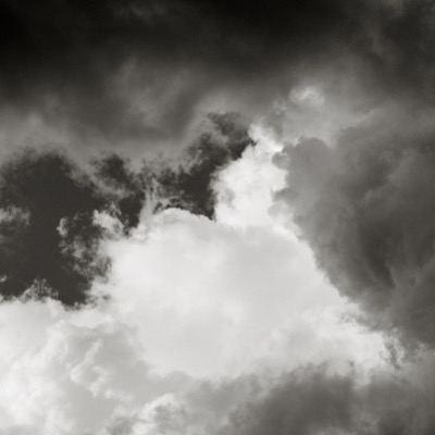 Black-and-white photograph of dense, dark cloud cover with a single luminous white cumulus formation glowing at its center.
